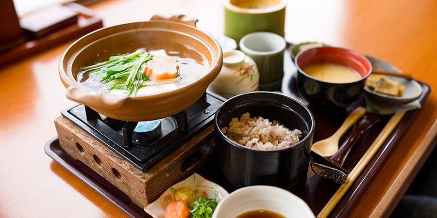 An assortment of small traditional Japanese dishes, including vegetables, garnishes and rice, and a bowl of miso soup being heated up by a small stove. All arranged neatly on a wooden tray on a wooden table.